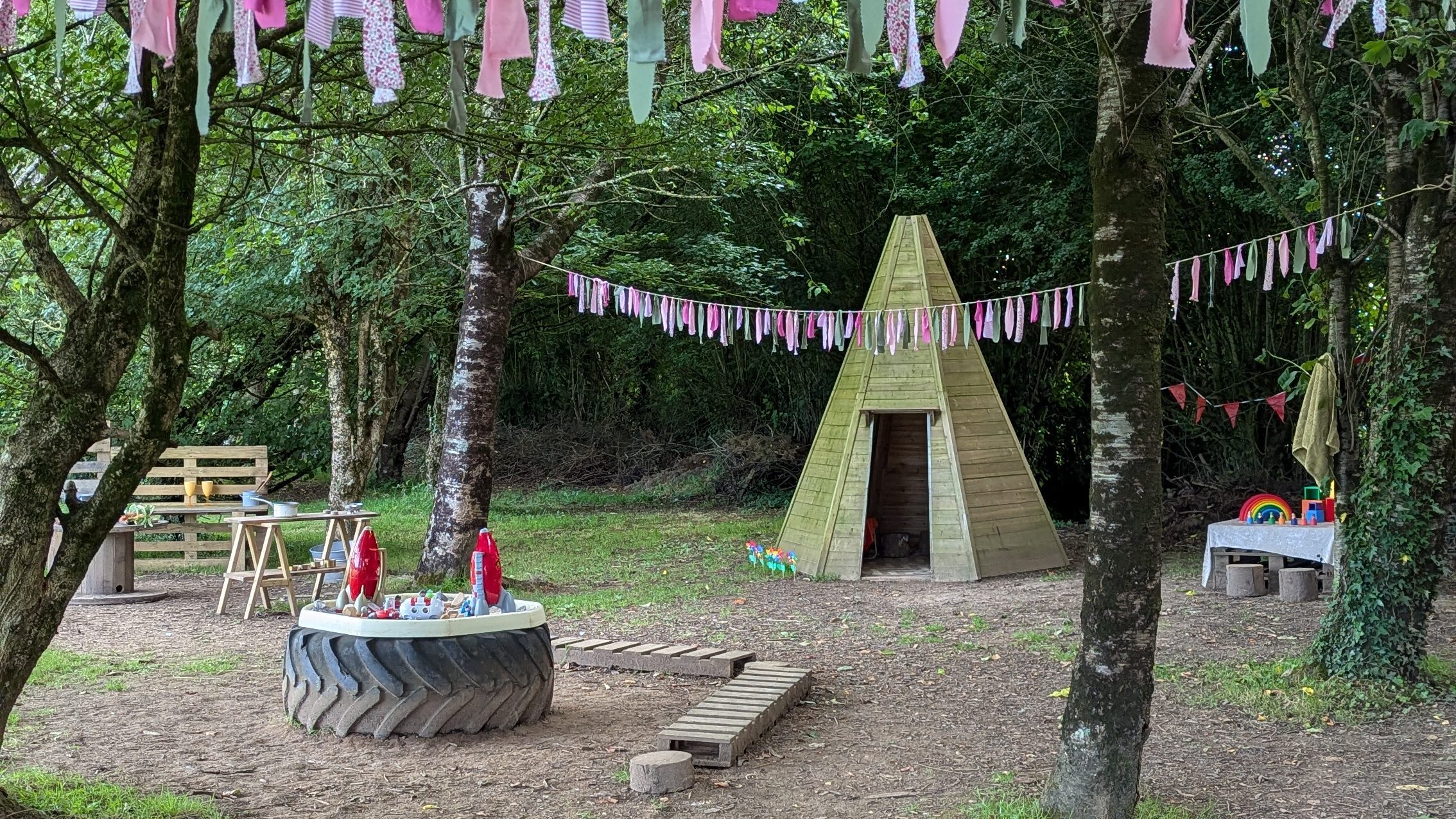 Photos shows our magical forest site in Bridgend at The Nature Play Patch. A tall teepee is surrounded by pink bunting, trees and leafy foliage. You can also see the other play stations set up - the mud kitchen, space station small world and colourful building blocks.